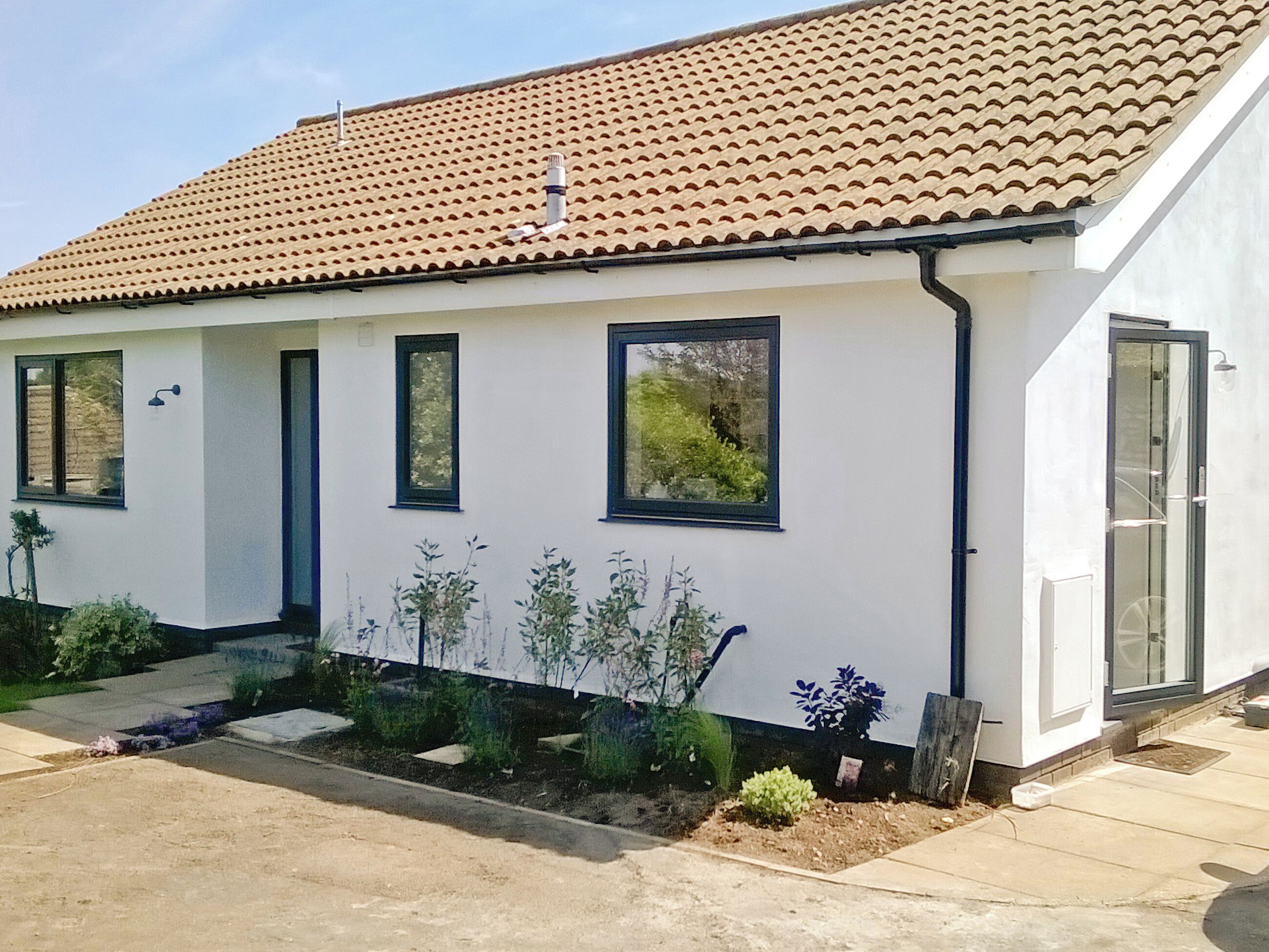 Eingeschossiges Haus mit Ziegeldach, weißen Außenwänden und einem kleinen Vorgarten mit verschiedenen Pflanzen.