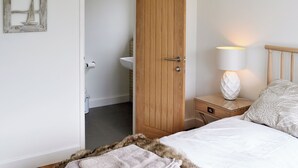 A bedroom with a wooden door, a bedside table with a lamp, a bed with a white duvet, and a small bathroom visible through the door. - 3 bedroom accommodation in Brancaster Staithe, near Hunstanton (Brancaster Staithe, near Hunstanton)