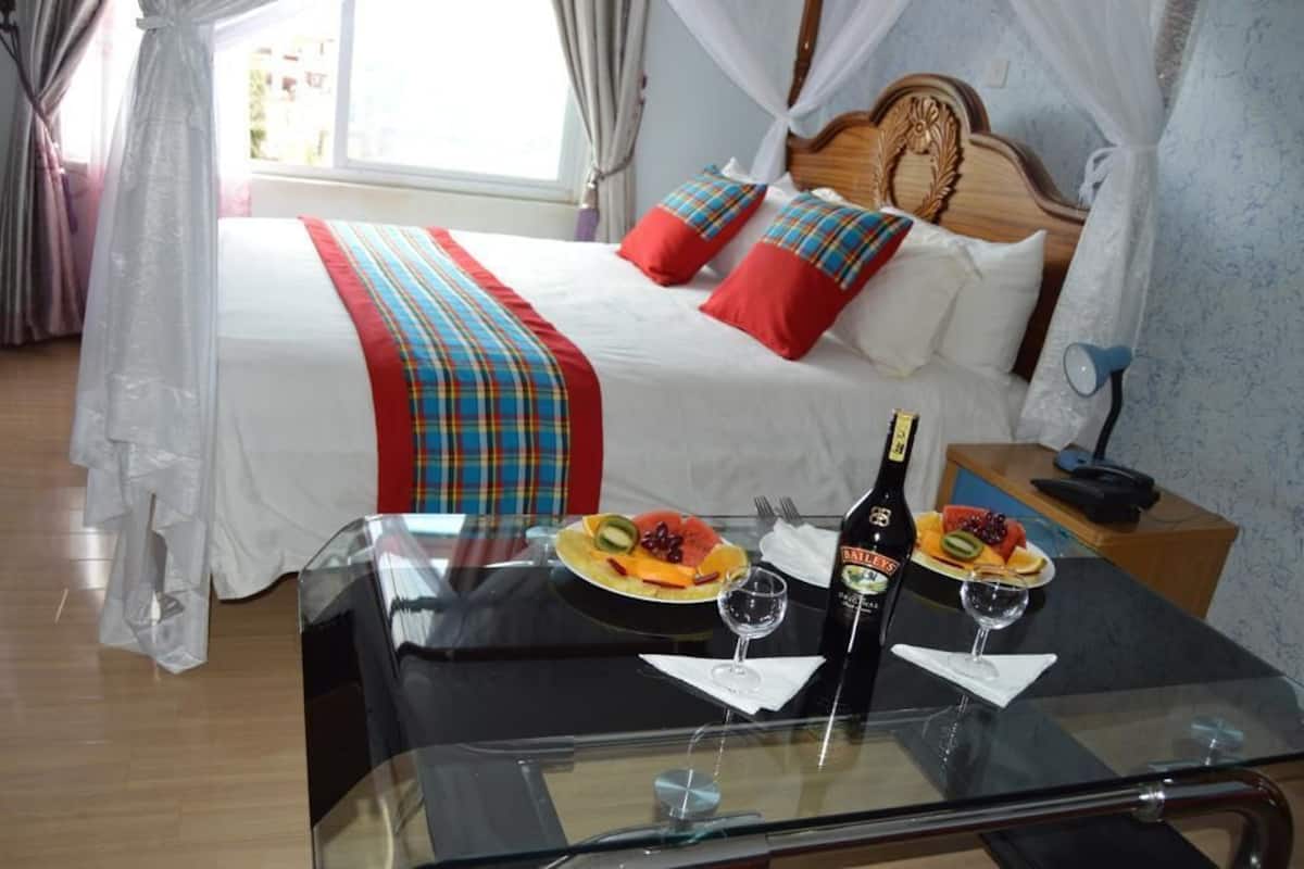 A hotel room with a bed, a glass table with breakfast, a bottle of wine, and a bedside table with a phone.