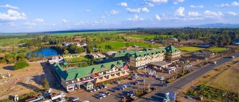 An aerial view of a hotel with a green-roofed building, a parking lot, and surrounding green fields and a lake.