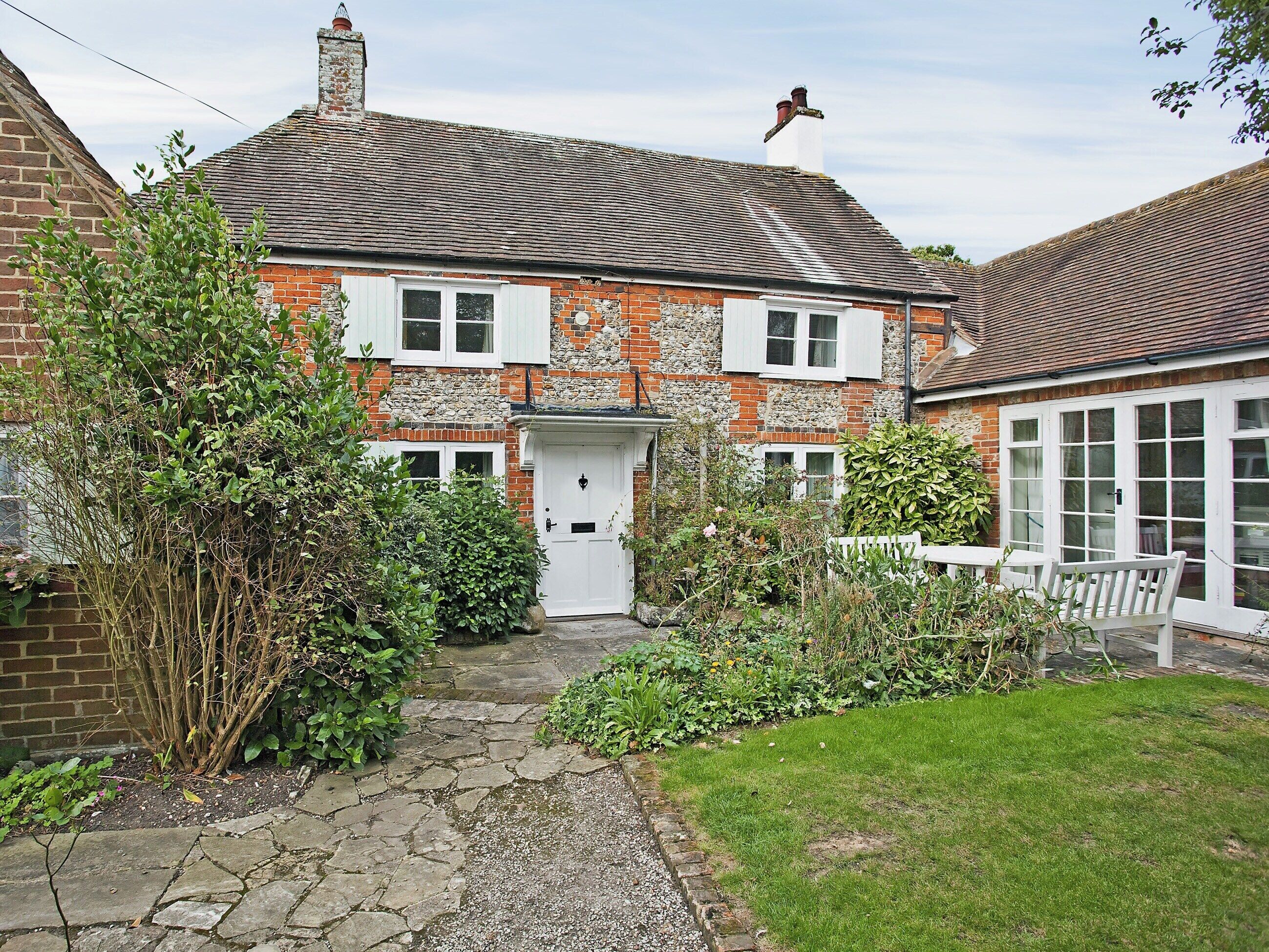 Une maison à deux étages avec une façade en pierre, une porte blanche et un petit jardin avec un chemin menant à l’entrée.