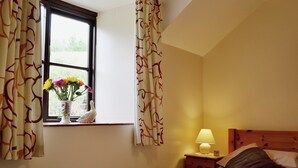 A bedroom with a bed, a nightstand with a lamp, a window with curtains, and a vase with flowers on the windowsill. - Honeysuckle Cottage - 29938 (Longdown, near Exeter)