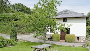 A white building with a stone foundation, a wooden door, and a small window. There is a wooden picnic table in the foreground with a grassy area around it. - Honeysuckle Cottage - 29938 (Longdown, near Exeter)