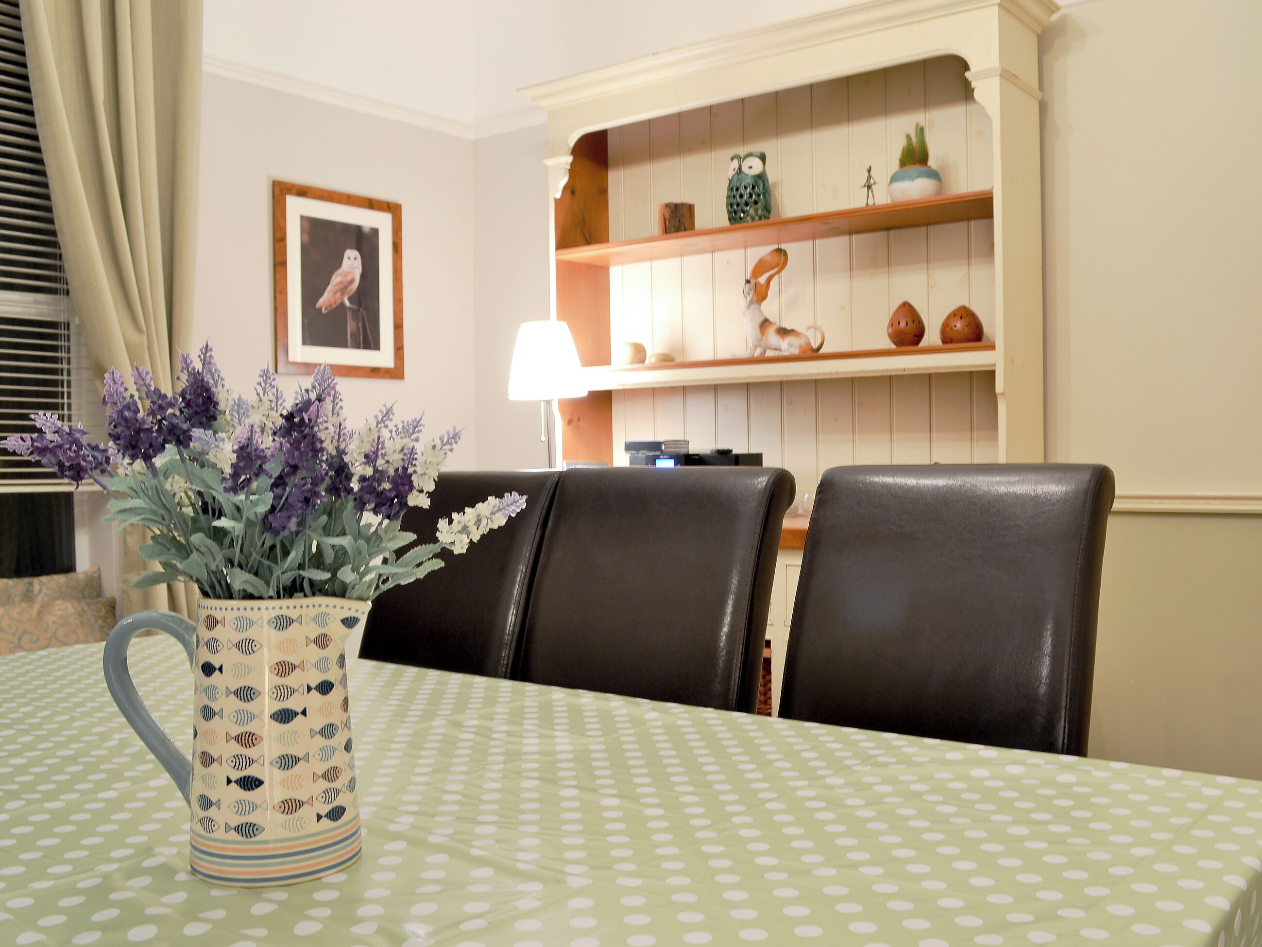 A dining area with a table covered by a green polka dot tablecloth, surrounded by black leather chairs. A vase with purple flowers is placed on the table.
