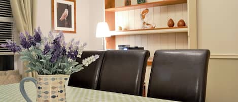 Ein Essbereich mit einem Tisch, bedeckt mit einer grün-weiß gepunkteten Tischdecke, umgeben von schwarzen Ledersesseln. Auf dem Tisch steht eine Vase mit lila Blumen.