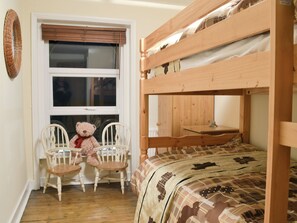 A bunk bed with a wooden headboard, a window with blinds, two white chairs, and a teddy bear.