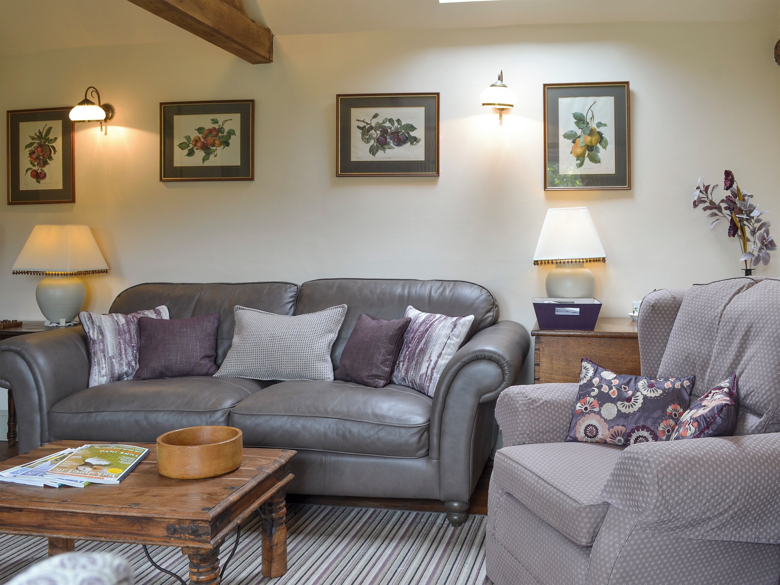A living room with a grey leather sofa, a wooden coffee table, and a patterned armchair. There are framed botanical prints on the wall and two table lamps.