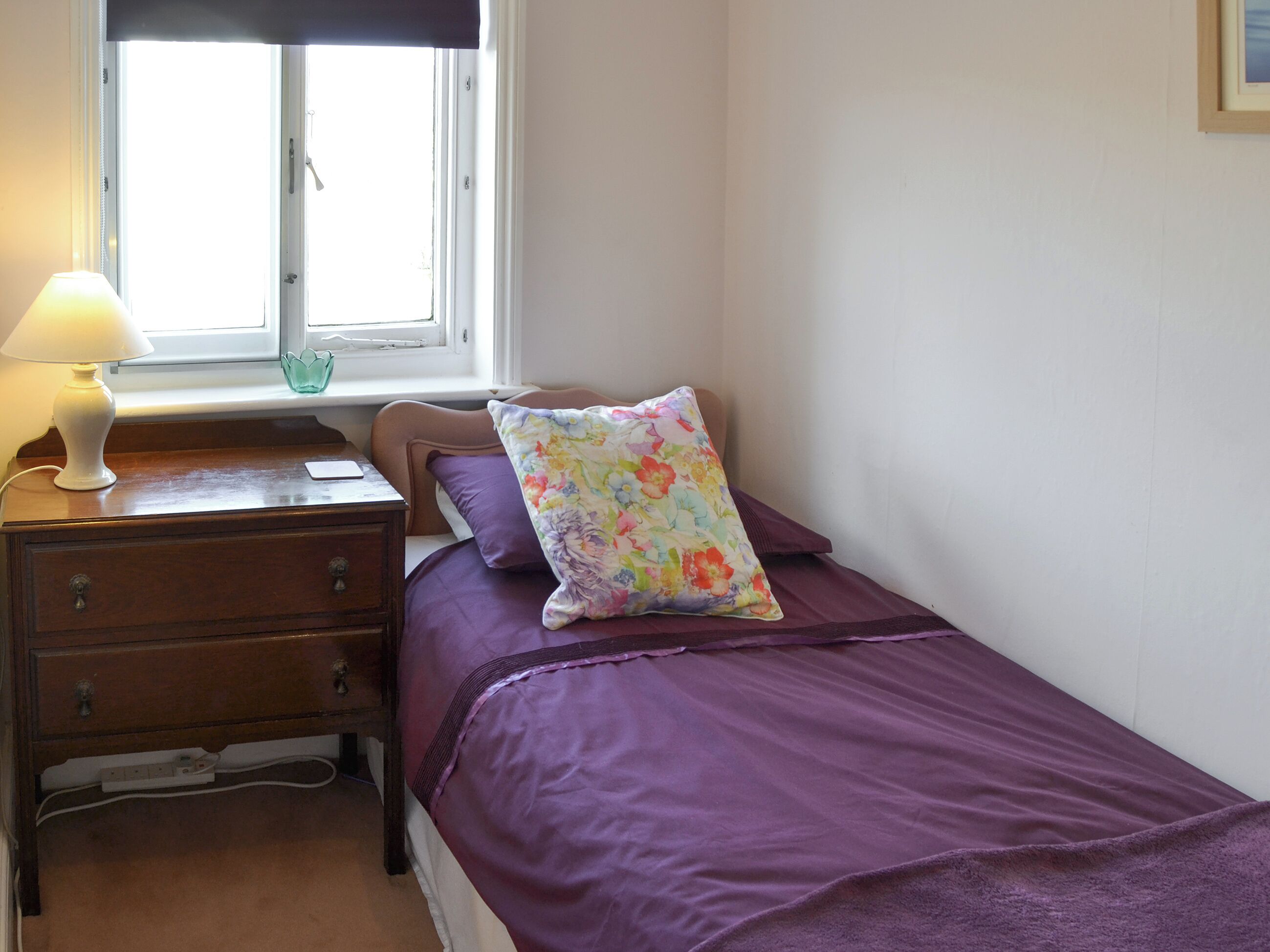 A bedroom with a bed, a nightstand, a lamp, a window, and a wall-mounted picture frame.