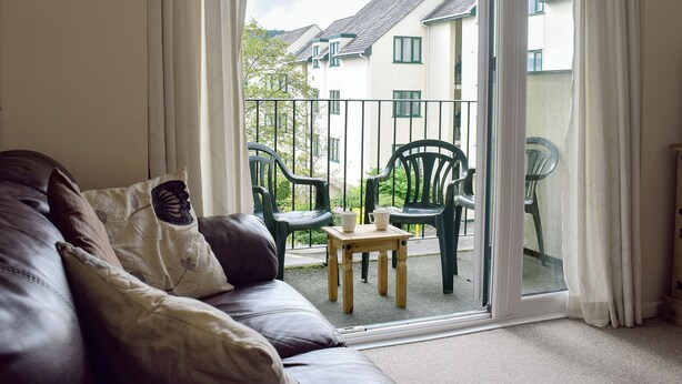 A living room with a leather sofa, a small wooden table, and two green chairs. A sliding glass door leads to an outdoor balcony with a metal railing and a view of buildings. - 2 bedroom accommodation in Bowness-on-Windermere (Bowness-on-Windermere)