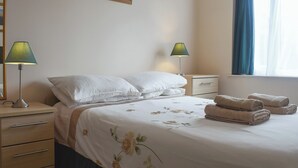A neatly made bed with white linens and floral patterns, two bedside lamps, a wooden nightstand, a drawer, and a window with blue curtains. - 2 bedroom accommodation in Bowness-on-Windermere (Bowness-on-Windermere)