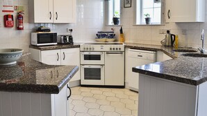 A modern kitchen with white cabinets, a microwave, a stove, and a dishwasher. There are two windows with blue curtains and a ceiling light fixture. - 4 bedroom accommodation in Wern-y-Wylan (Wern-y-Wylan)