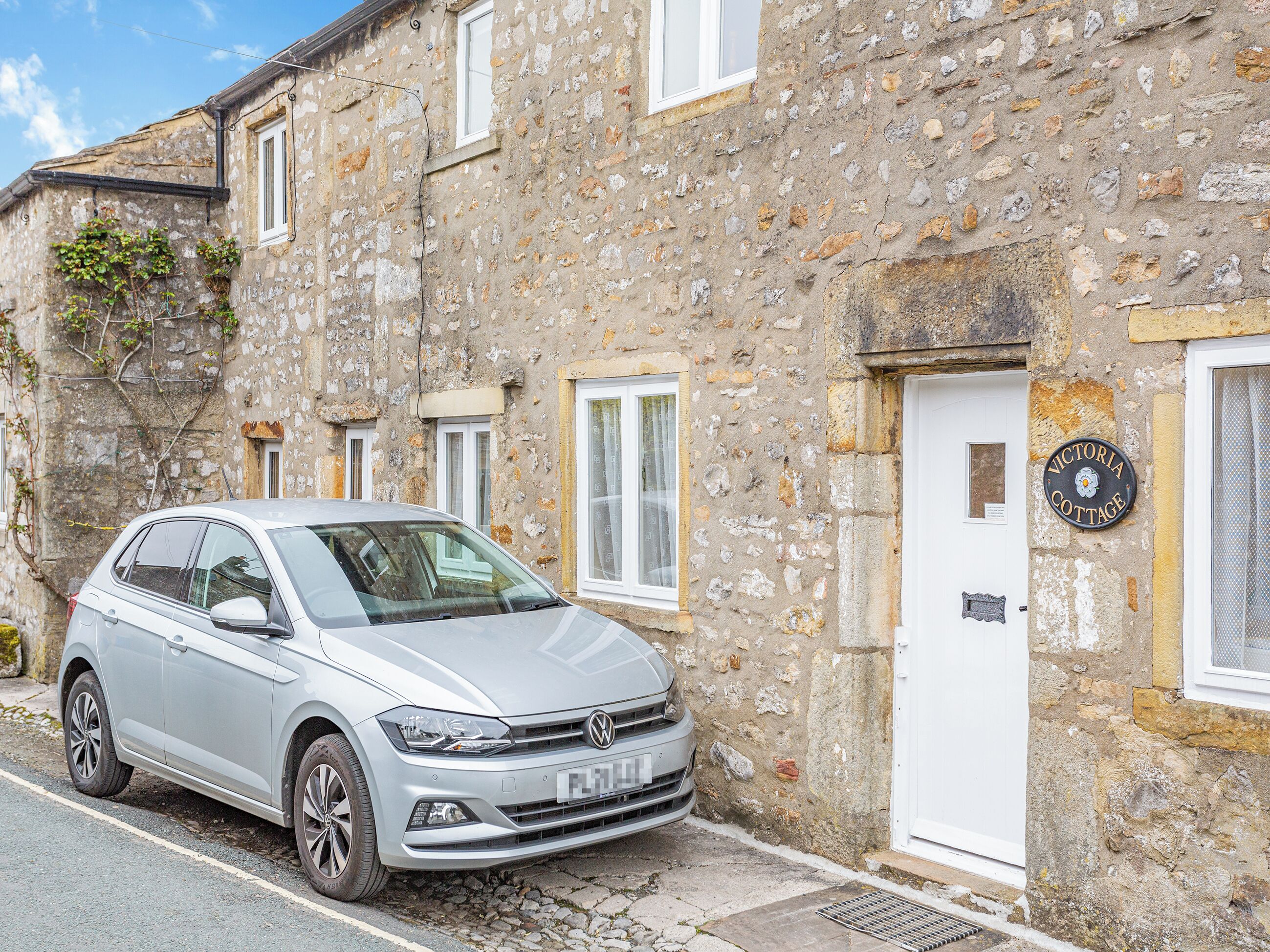 A silver Volkswagen car parked in front of a stone building with a white door and a sign that reads "Victoria Cottage."