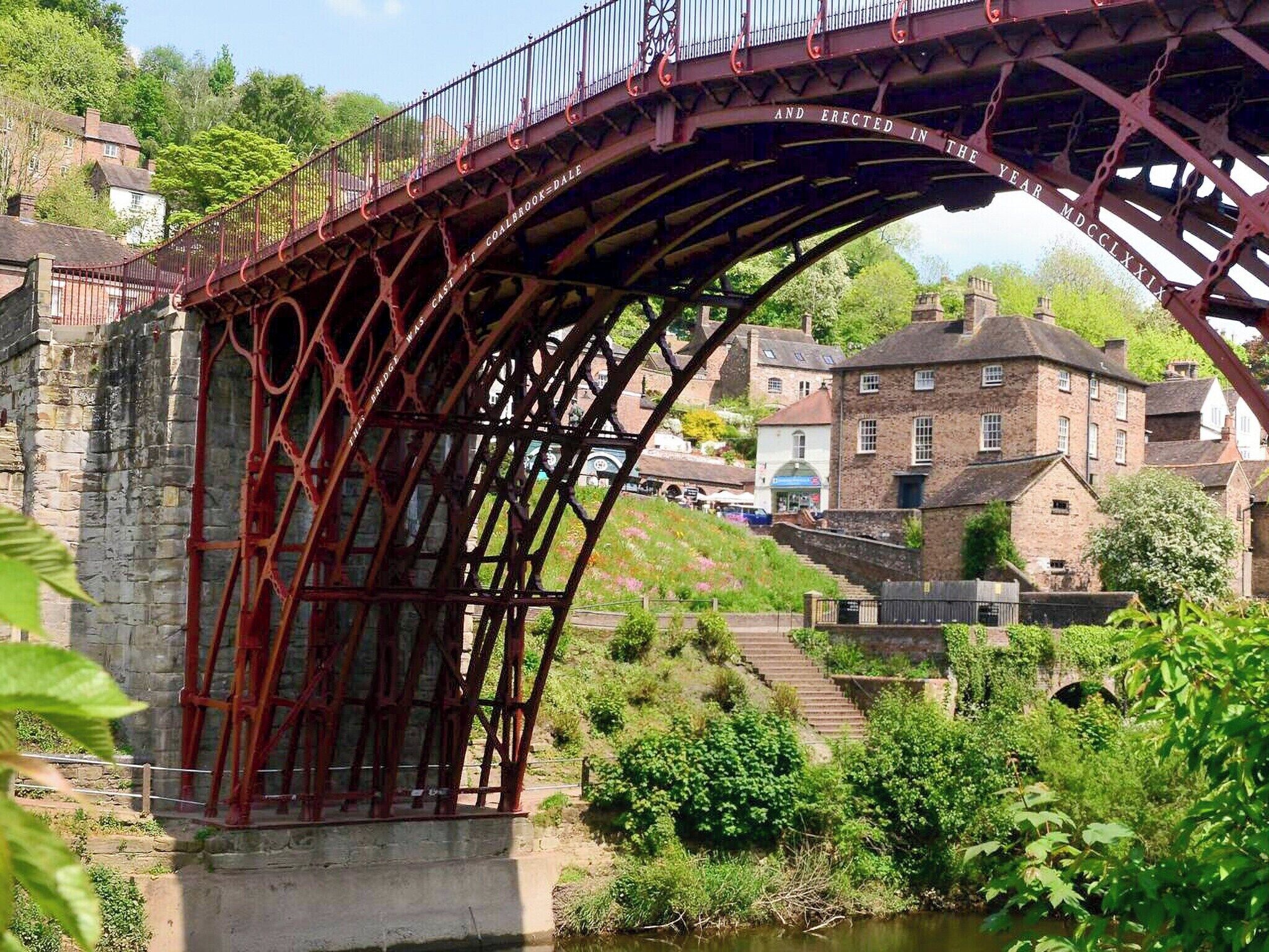 Un ponte in metallo rosso con iscrizioni, un muro in pietra e un edificio con tetto a forte pendenza.