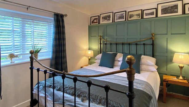 Een netjes opgemaakt slaapkamer met een bed, nachtlampjes, een raam met zonwering en een muur met ingelijste foto’s.