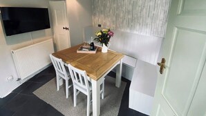Een eethoek met een houten tafel, witte stoelen, een aan de muur bevestigde televisie en een koelkast.