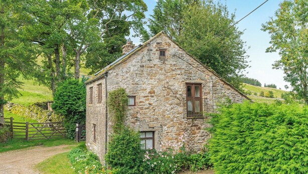 Una casa de piedra con techo inclinado, rodeada de abundante vegetación y un sendero de grava que lleva a un portón de madera.