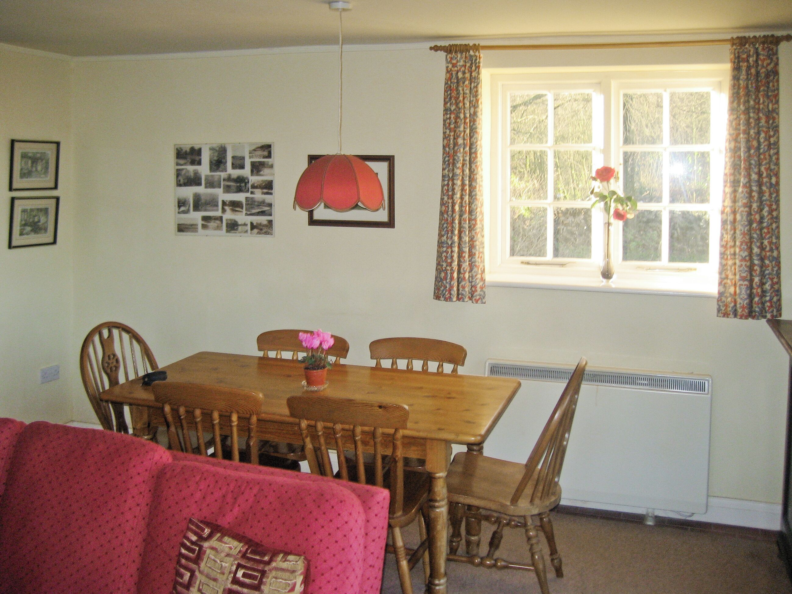 A dining room with a wooden table and chairs, a red pendant light, a window with curtains, and a radiator.