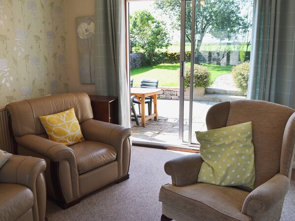 A living room with two leather armchairs, one with a yellow pillow and the other with a green pillow. A wooden table and chairs are visible through a glass door leading to an outdoor area with a lawn and a stone wall.
