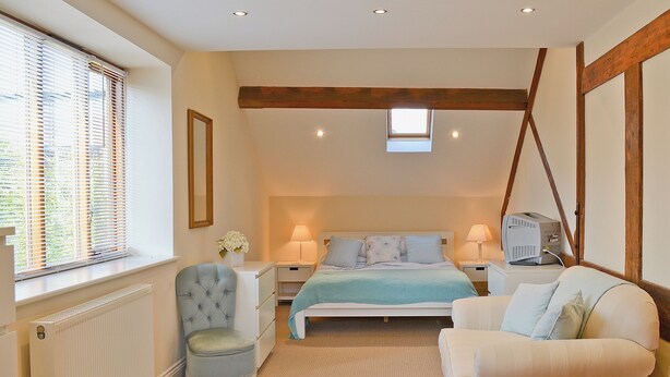 A bedroom with a bed, two bedside tables, a chair, a sofa, a radiator, a window with blinds, a skylight, and wooden beams on the ceiling. - 4 bedroom accommodation in Alvechurch (Alvechurch)
