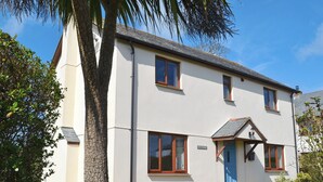 A two-story house with a blue door and windows, surrounded by a lawn and trees. - 4 bedroom accommodation in Marhamchurch, near Bude (Marhamchurch, near Bude)