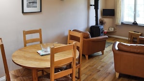 Een gezellige woonkamer met een ronde houten tafel, twee houten stoelen, een bruine leren bank, een open haard en een raam met gordijnen.