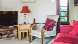 A living room with a television, a red lamp, a chair with a cushion, and a red sofa. - 2 bedroom accommodation in Buckfastleigh (Buckfastleigh)