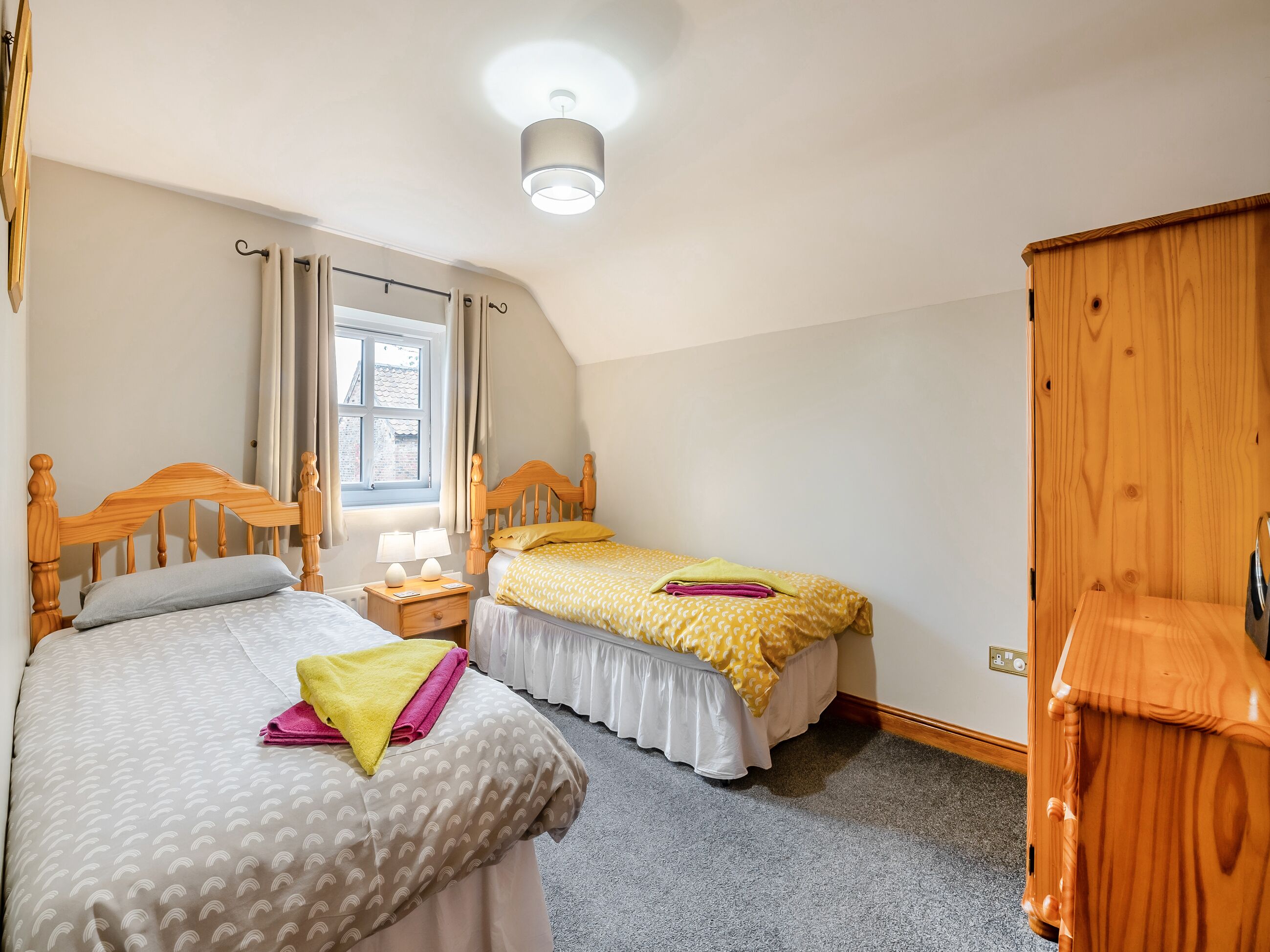 Une chambre à coucher avec deux lits en bois, une table de chevet avec des lampes, une fenêtre avec des rideaux et une armoire en bois.