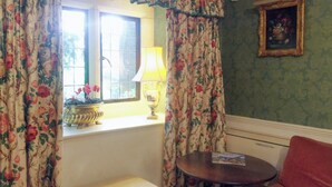 A room with floral curtains, a window with a view, a table with a lamp, and a framed picture on the wall.