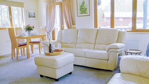 Una sala de estar con un sofá blanco, un reposapiés a juego, una mesa de comedor de madera con sillas y una ventana con cortinas.
