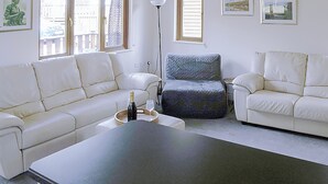 Una sala de estar con dos sofás blancos de cuero, un sillón gris, una mesa de centro y una lámpara de pie. Hay cuadros enmarcados en la pared y una ventana con persianas.