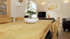 A dining area with a wooden table, chairs, a vase with flowers, a television, and a sofa in the background.