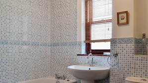 Une salle de bain équipée d’une baignoire, d’un lavabo et de toilettes, avec des carreaux bleus à motifs et une fenêtre avec des stores.