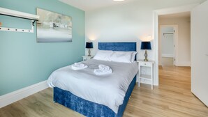 A bedroom with a bed, two bedside tables, a nightstand, a wall-mounted lamp, a painting, and a door leading to another room. - Silverdale Apartment (Meathop, near Grange-over-Sands)