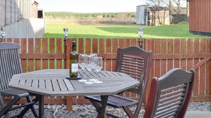 A wooden table with a bottle of wine and glasses, set against a wooden fence with a view of a grassy field and buildings in the background.
