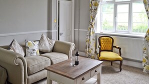 A living room with a beige sofa, a yellow armchair, a wooden coffee table, and a window with floral curtains.