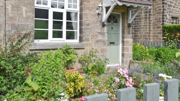 A stone house with a white window and door, surrounded by a garden with various flowers and plants.