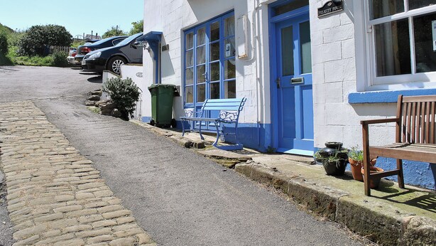 Un bâtiment blanc avec des bordures bleues et une porte bleue, un banc et une chaise en bois à l’extérieur.
