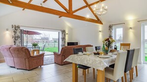 A spacious living area with a high ceiling and exposed wooden beams, a dining table set for breakfast, and a view through large glass doors to the outdoors. - 3 bedroom accommodation in Wicken, near Ely (Wicken, near Ely)