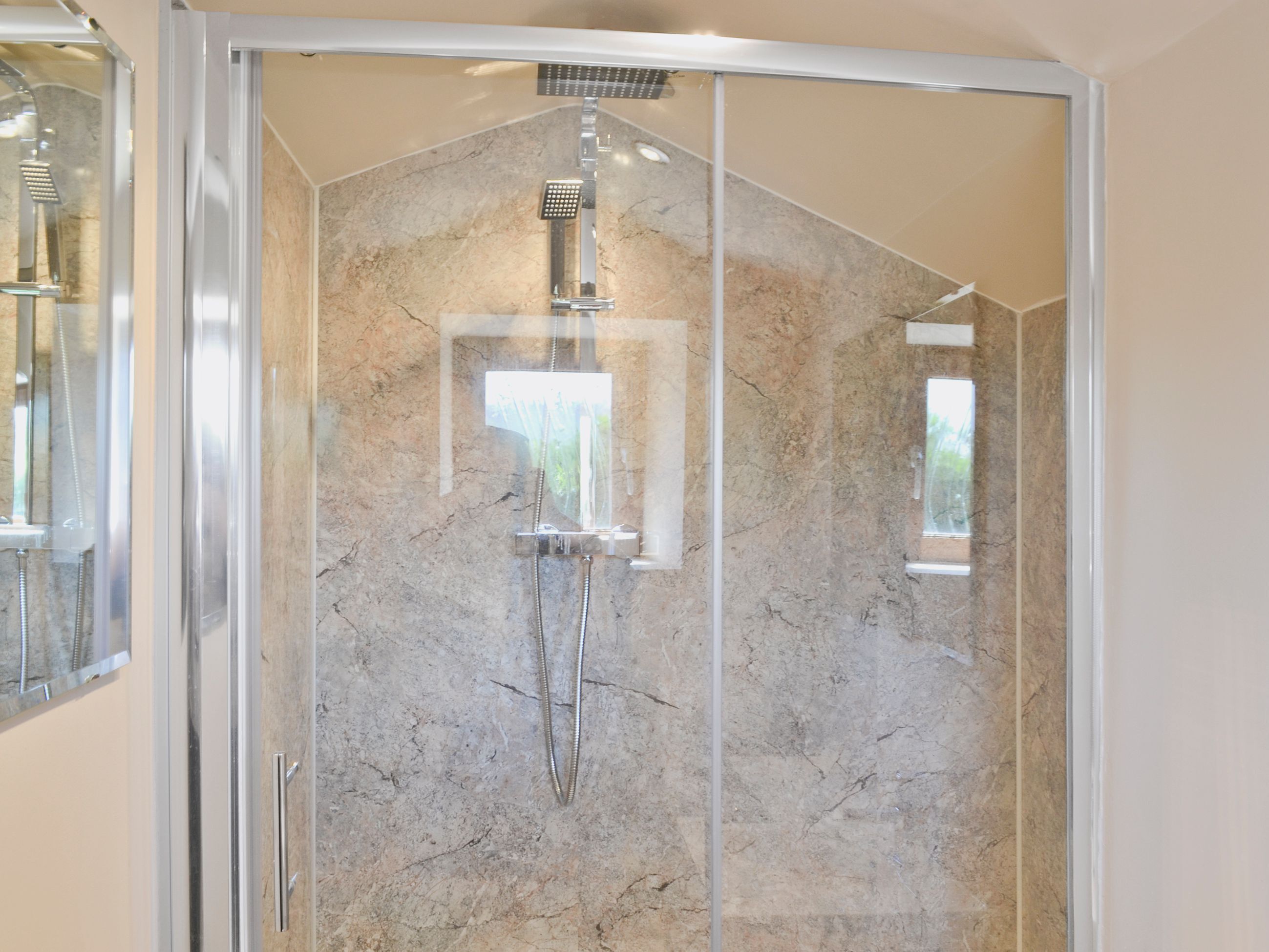 A glass-enclosed shower with a marble wall and a handheld showerhead.