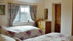 A bedroom with two beds, a wooden dresser, a window with floral curtains, and a mirror on the wall.