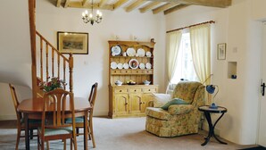A cozy living room with a wooden dining table, chairs, a floral armchair, a side table with a lamp, and a china cabinet.