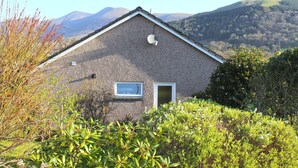 A house with a sloped roof, a small window, and a door. It is surrounded by greenery and hills in the background.