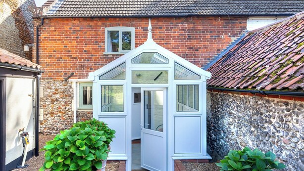A small white greenhouse with a door, flanked by two large potted plants, set against a brick wall and a tiled roof. - 2 bedroom accommodation in Docking, near Hunstanton (Docking, near Hunstanton)