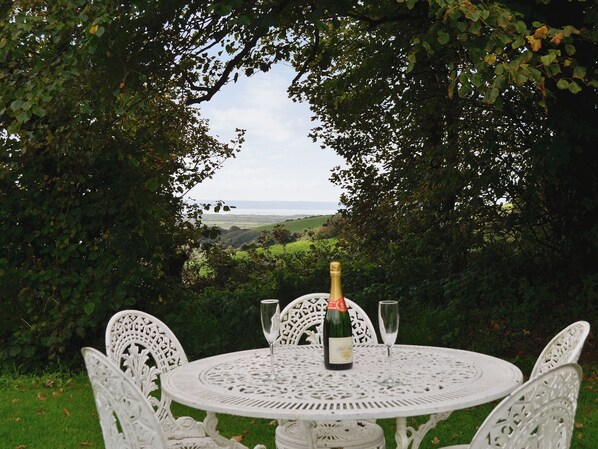 Une table ronde avec une bouteille et deux verres, entourée de chaises blanches, installée à l’extérieur avec vue sur la mer.