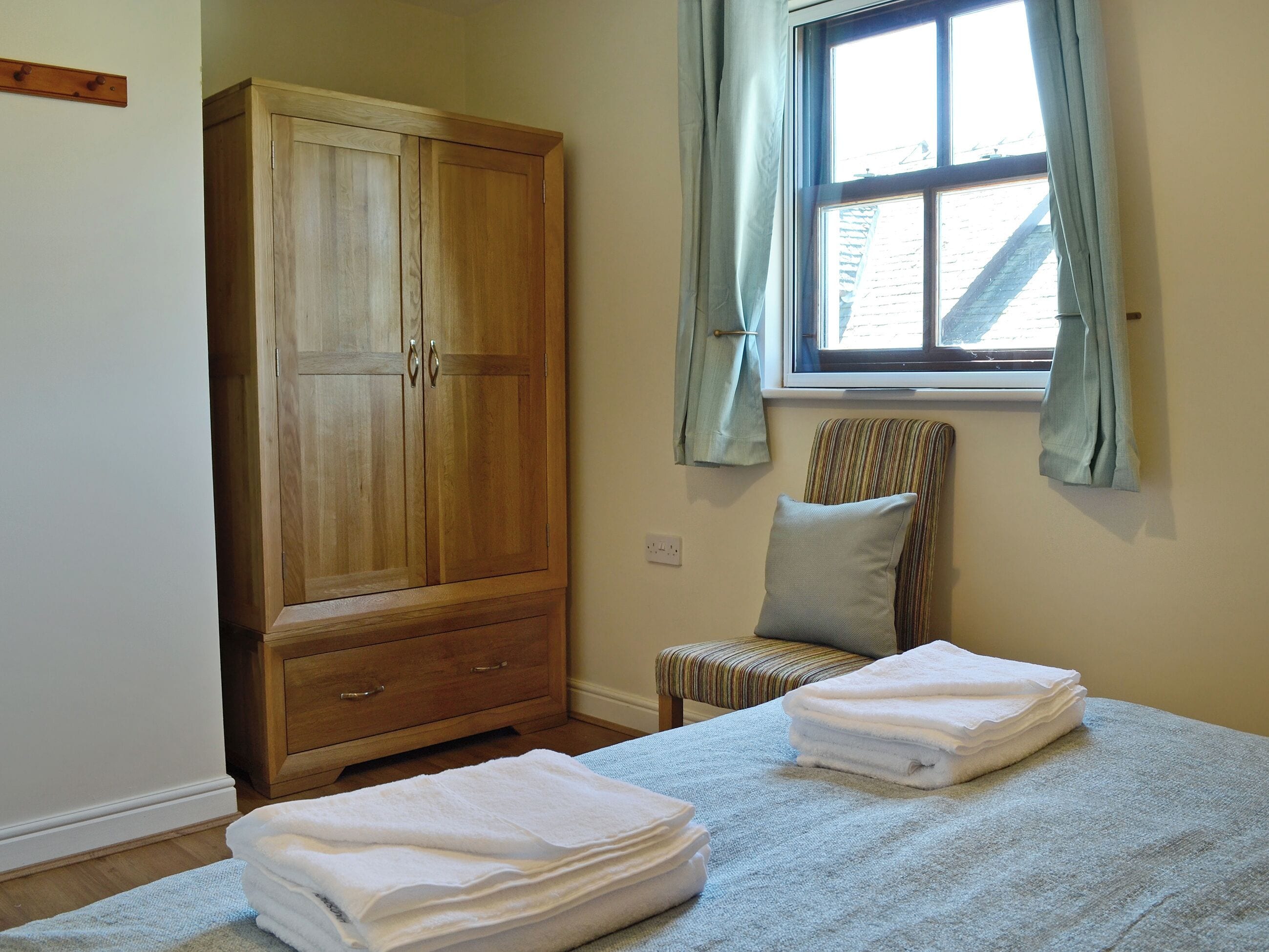 Ein Schlafzimmer mit einem Holzschrank, einem Fenster mit Vorhängen, einem gestreiften Sessel und ordentlich gefalteten Handtüchern auf dem Bett.