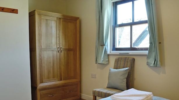 Une chambre avec une armoire en bois, une fenêtre avec des rideaux, une chaise à rayures et des serviettes soigneusement pliées sur le lit.