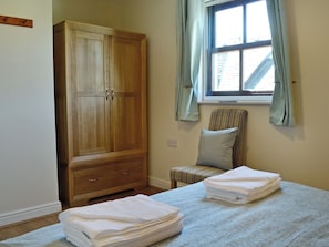 A bedroom with a wooden wardrobe, a window with curtains, a striped chair, and neatly folded towels on the bed.