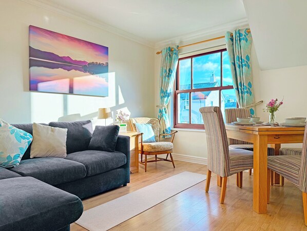 A living room with a sofa, a dining table with chairs, a window with curtains, and a painting on the wall.