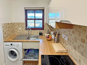 A compact kitchen with a washing machine, a sink, a window with a view, and a countertop with a bowl of fruit.
