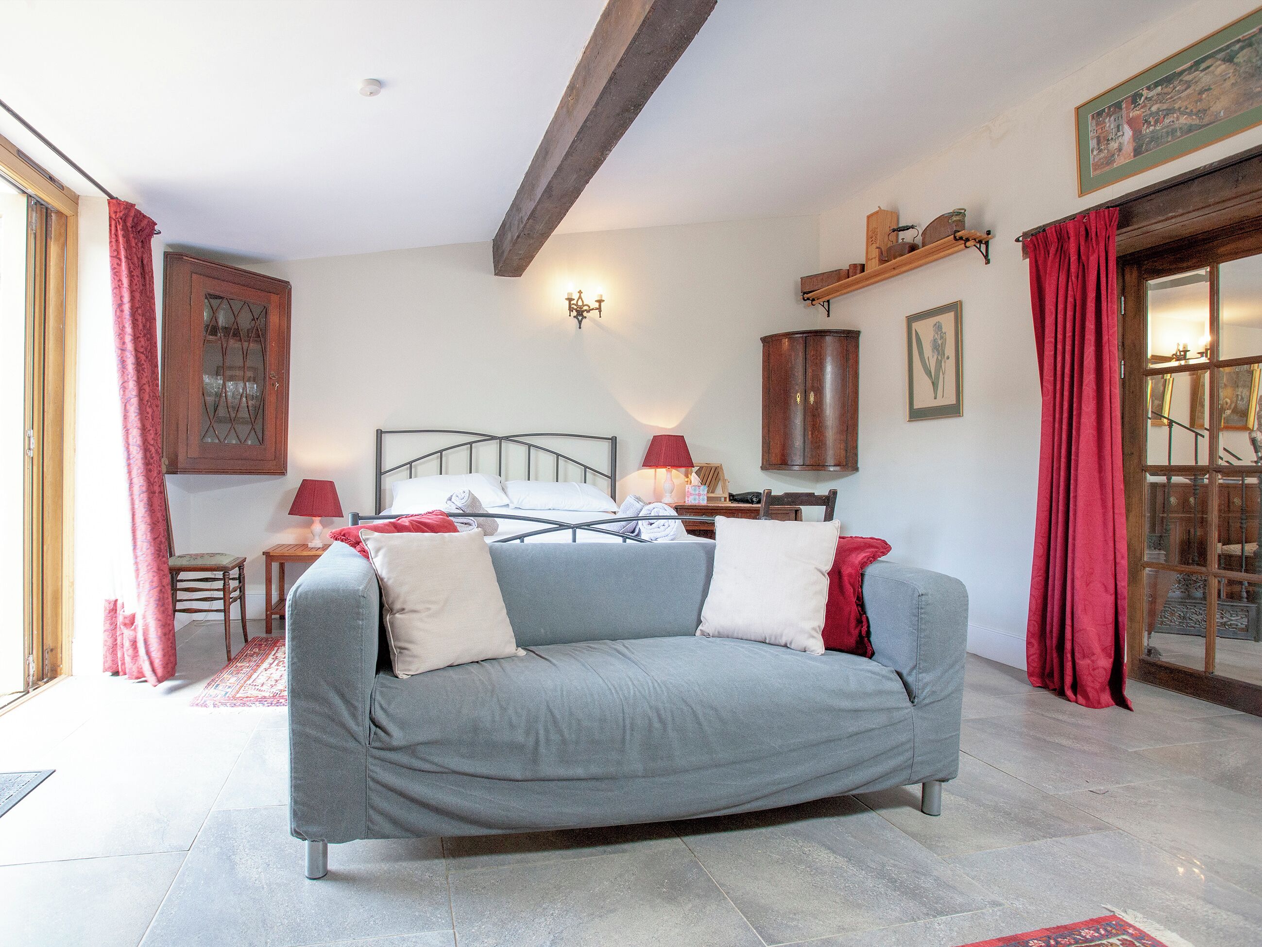 Quarto com sofá cinza, um armário de madeira, cortina vermelha e cama com roupa de cama branca.